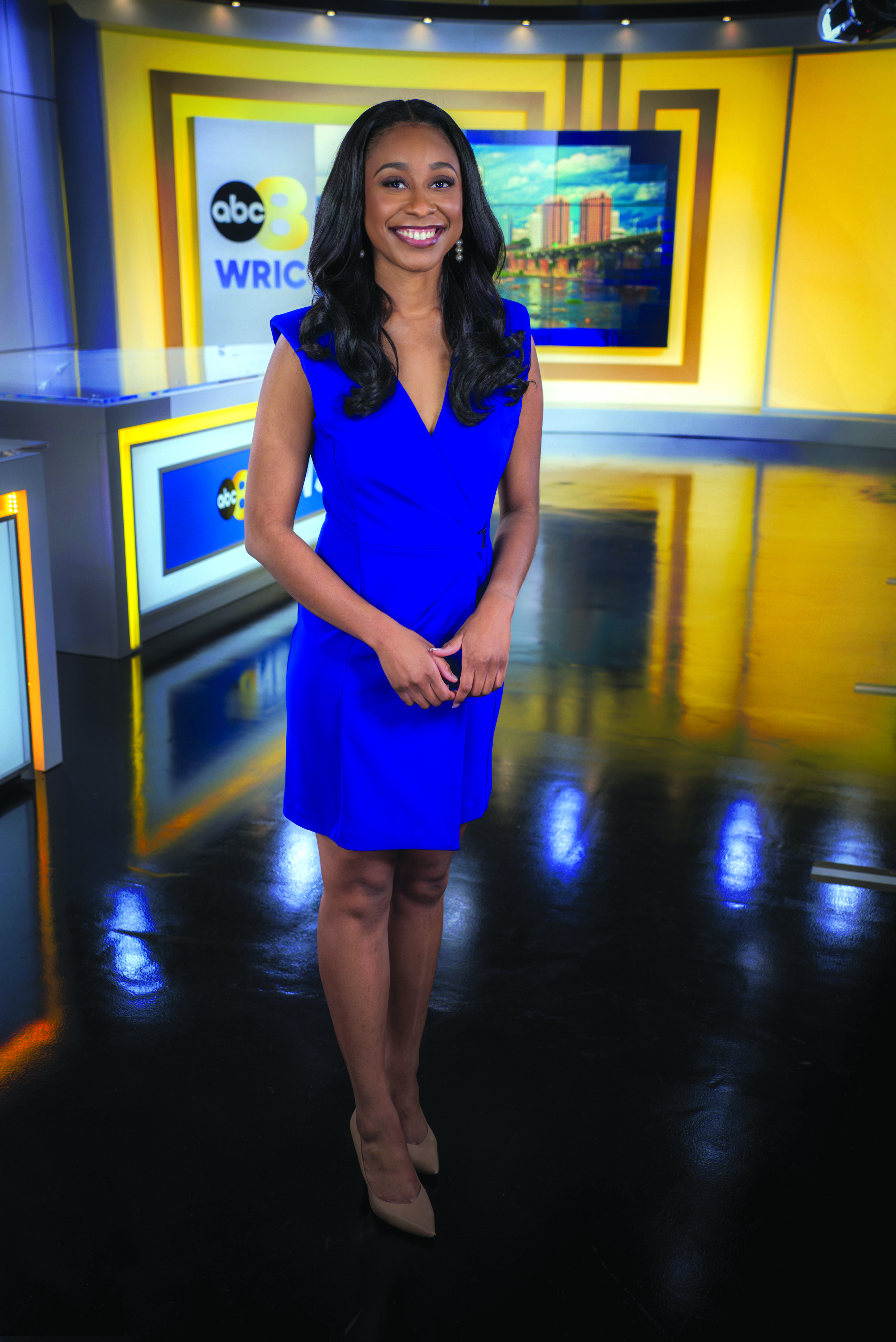 A woman stands in a television news studio, smiling confidently at the camera. She is wearing a blue dress and beige high heels. The background features bright yellow accents, a news desk, and a screen displaying the logo 