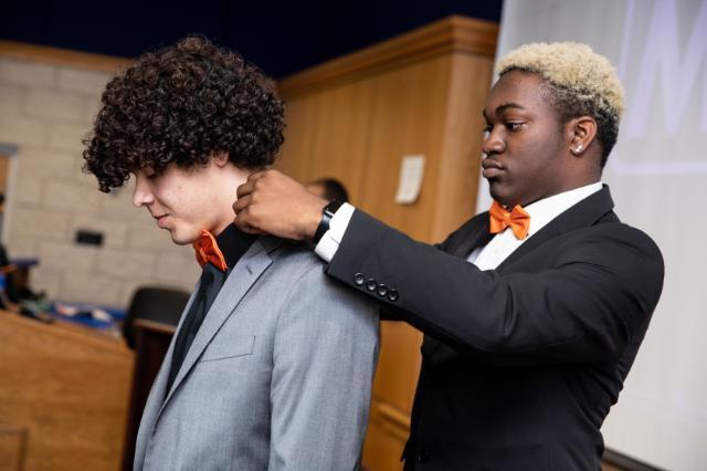 A young man in a black suit and orange bow tie helps adjust the bow tie of another participant wearing a gray suit during a Call Me MiSTER ceremony.