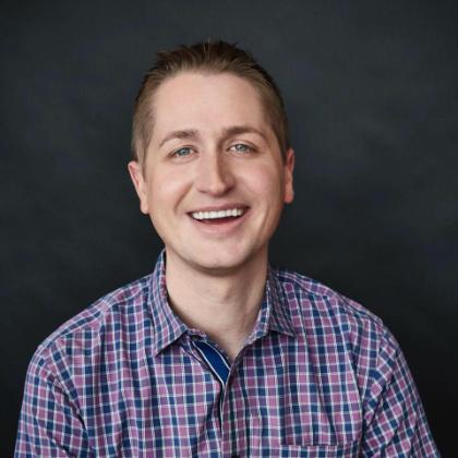 Smiling man in a purple and blue checkered shirt against a dark background.