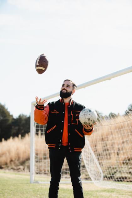 A man standing on a grassy field in front of a soccer goal, wearing a black and orange varsity jacket with a large letter 